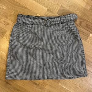 Kortkjol med bälte - Snygg rutig kortkjol med svartvita små rutor (kjolen upplevs som grå) och ett matchande bälte med rund metallspänne. Kjolen är använd max fem gånger. Storlek L/40.