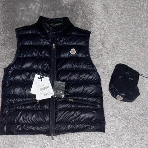Svart dunväst från Moncler - Snygg blå dunväst från Moncler med klassisk quiltad design och dragkedja framtill. Västen har ståkrage, två fickor med dragkedja och Moncler-logga på bröstet, västen är storlek 4 vilket motsvarar ca large!