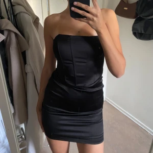 Svart axelbandslös kort klänning - Säljer en svart, axelbandslös kort klänning. Klänningen har en snygg, korsettliknande överdel och är sydd i siden material. 