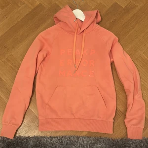 Korall hoodie från Peak Performance XS - Säljer en snygg korallfärgad hoodie från Peak Performance i storlek XS. Tröjan har huva med snörning, stor magficka och tryckt logga framtill. Perfekt för chill dagar eller när du vill ha en färgklick i garderoben.