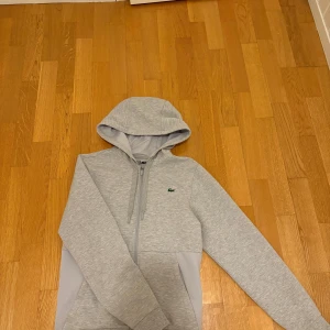Grå hoodie från Lacoste Sport XS - Snygg ljusgrå hoodie från Lacoste Sport i storlek XS. Tröjan har dragkedja, huva med snörning och klassisk krokodillogga på bröstet. Tillverkad i mjuk bomullsblandning med mesh-detaljer på sidorna för extra komfort. Perfekt för chill eller träning.