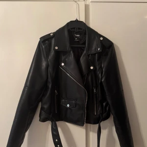 Svart skinnjacka från Pull&Bear XL - Snygg svart skinnjacka från Pull&Bear i klassisk bikerstil. Jackan har silvriga dragkedjor, knappar och bälte i midjan. Modellen är croppad med långa ärmar och coola detaljer som axelklaffar och fickor framtill. Den är sönder i hängöglan men annars helt perfekt. Jag står ej för frakten!