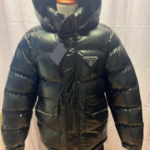 Svart pufferjacka från Prada - Snygg svart pufferjacka från Prada med glansig finish och stor huva. Jackan har två stora fickor framtill, dragkedja och tryckknappar. Klassisk Prada-logga på bröstet och cool, oversized look. Perfekt för kalla dagar när du vill se extra fresh ut.