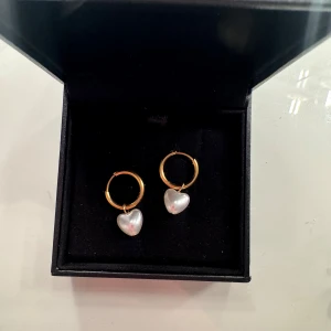 Guldiga örhängen med pärlhjärtan - Supersöta örhängen med guldfärgade hoops och hängande vita pärlhjärtan. Perfekta för att ge din look en romantisk touch! Hjärtformade detaljer gör dem extra unika och de kommer i en snygg svart ask.