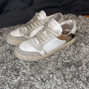 Burberry vita sneakers med rutiga detaljer - Snygga vita sneakers från Burberry med klassiska rutiga paneler på sidan. (Kalvskin) Skorna har snörning, platt sula och rund tå. Materialmix av skinn och mocka med beige och grå toner. Perfekt för dig som gillar designer och vill sticka ut med en ikonisk detalj. Skinet har spruckit på vissa ställen(se bilderna)