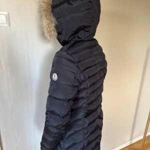 Svart Moncler dunjacka med pälskant - Säljer en svart lång dunjacka från Moncler med huva och snygg pälskant. Jackan har quiltad design, dragkedja och Moncler-logga på ärmen. Perfekt för kalla dagar när du vill hålla stilen och värmen.