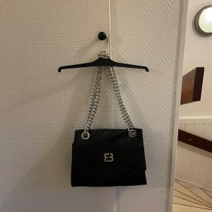 Svart axelväska med kedja från Fanny Blomme - Snygg svart axelväska med grov silverkedja som axelrem och metalliskt FB-logo framtill. Väskan har quiltad yta och är tillverkad i mjukt syntetmaterial. Perfekt storlek för det viktigaste och ger en cool vibe till din outfit.