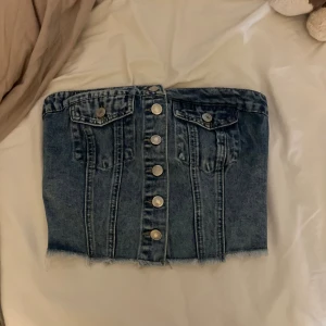 off shoulder topp - Säljer en off shoulder topp i blå denim med fransad nederkant och silverfärgade knappar framtill. Kjolen har två fickor med lock och knapp på framsidan samt smockad detalj baktill för extra stretch. Perfekt för dig som gillar en cool och avslappnad stil.