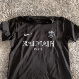 Svart PSG x Balmain fotbollströja - Snygg svart fotbollströja från Nike i samarbete med Balmain och Paris Saint-Germain. Tröjan har korta ärmar, rund hals och tryck i vitt framtill med både Balmain och PSG-logga. Perfekt för dig som gillar exklusiva samarbeten och sportig stil.  Detta är en Replika!