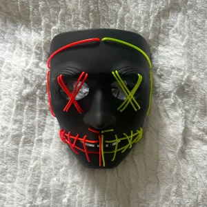 Svart LED-mask med neonröd och grön - Cool svart mask med LED-linjer i neonrött och neongrönt som lyser upp i mörkret. Masken har X över ögonen och ett rutmönster över munnen, perfekt för att sticka ut på festen eller temakvällen. Drivs med batteridosa. Har tre olika läggen vanlig lyssa hela tiden, blinkade och blinka snabbt.  Endast använd en kväll 