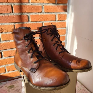 Rieker skinn Kängor, Brun, Storlek 42 - Snygga bruna kängor från Rieker med snörning och dragkedja på sidan. Tillverkade i skinn med detaljer i mörkbrunt och metallknappar. Insidan är fodrad med grått ullmaterial för extra värme. Rund tå och platt sula ger en klassisk look.