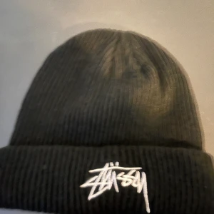 Svart Stüssy mössa med broderad logga - Svart ribbstickad mössa från Stüssy med vit broderad logotyp framtill. Klassisk streetwear-stil med uppvikt kant, perfekt för kalla dagar. Materialet är mjukt och stretchigt för skön passform.