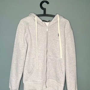 Grå hoodie från Polo Ralph Lauren - Ljusgrå hoodie från Polo Ralph Lauren med dragkedja, huva och vita snören. Klassisk broderad logga på bröstet och två fickor framtill. Perfekt för chill dagar eller när du vill ha en clean och enkel look.