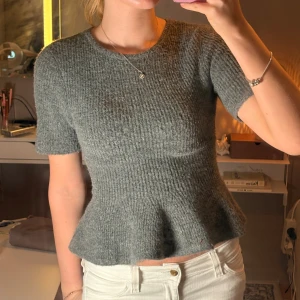 Grå stickad topp med volangkant  - Superfin grå stickad topp med korta ärmar och volangkant nedtill från HM! JÄTTE populär och trendig just nu. Höstens trend!! Toppen har en rund halsringning och är figurnära upptill med en lösare passform vid midjan. Perfekt för dig som gillar en enkel men ändå söt stil. Knappast använd