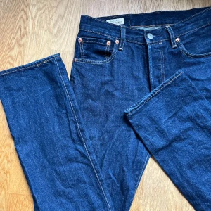 Levi's 501 mörkblå jeans straight fit - Klassiska Levi's 501 jeans i mörkblå denim med raka ben och normal passform. Byxorna har fem fickor, knappar i gylfen och tydliga kontrastsömmar. Ikonisk läderpatch bak och röd Levi's-tagg på bakfickan. Perfekta för en tidlös look. Pris kan diskuteras 