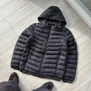 Svart pufferjacka från Stone Island - Säljer en svart pufferjacka från Stone Island med huva och dragkedja framtill. Jackan har det klassiska Stone Island-märket på ärmen och är quiltad för extra värme. Perfekt för kyliga dagar och har en clean, modern look.