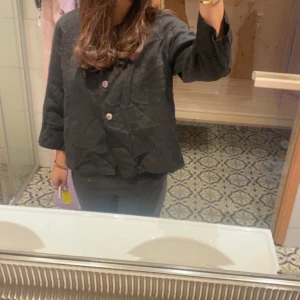 Svart linnekofta från Adini - Säljer en svart kofta från Adini i linne med tre stora knappar framtill. Koftan har en lös passform, rundad halsringning och trekvartsärmar. Perfekt för lager-på-lager och chill stil. Passar dig som gillar minimalistiska och stilrena plagg.