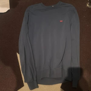 Blå sweatshirt från Levi's, storlek M - En stilren blå sweatshirt från Levi's med klassisk rund hals och liten röd logotyp på bröstet. Tröjan har långa ärmar och ribbade muddar vid ärmslut och nederkant. Perfekt för en avslappnad och snygg look.