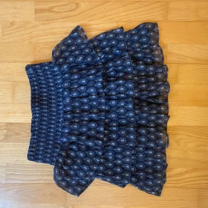 Blå volangkjol med mönster - Supersöt blå kortkjol med volanger och elastisk midja. Kjolen har ett småmönstrat tryck i vitt och blått och är perfekt för dig som gillar en lekfull och trendig stil. Lätt och luftig, passar perfekt till sneakers eller sandaler.