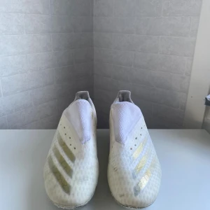 Vita och guldiga adidas fotbollsskor - Säljer ett par vita fotbollsskor från adidas med guldiga detaljer och klassiska tre ränder på sidan. Skorna har låg profil, plös utan snörning och ovandel i syntetmaterial. Perfekta för dig som vill sticka ut på planen med stilrena och sportiga skor. Har inga skdor men har tecken på använding.