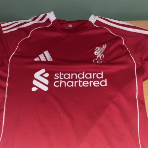 Liverpool FC röd matchtröja Adidas L - Liverpool FCs officiella matchtröja i rött med vita detaljer och klassiska Adidas-ränder på axlarna. Tröjan har klubbmärke, sponsorer och är tillverkad i ett lätt, ventilerande material. Slim fit-modell med korta ärmar och rund hals.