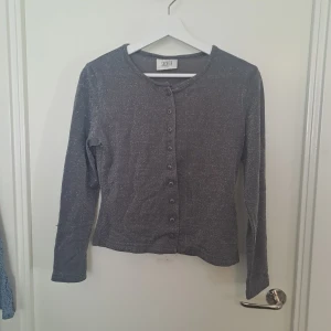 Glittrig grå långärmad topp - Grå långärmad topp från Suchita med glittrigt lurex-tyg och rund hals. Knappar framtill. Materialet är 83% nylon och 17%. 🩷