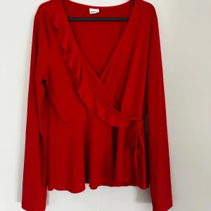 Röd omlottopp med volang från Gina Tricot - Säljer en snygg röd omlottopp från Gina Tricot med långa ärmar och volangdetalj längs v-ringningen. Toppen har knyt i sidan