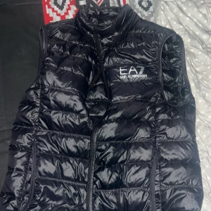 Svart dunväst från Emporio Armani EA7 - Snygg svart dunväst från Emporio Armani EA7 med glansig finish och horisontella quiltade sömmar. Västen har dragkedja framtill och logotyp på bröstet. Perfekt att slänga över en hoodie för en cool look.