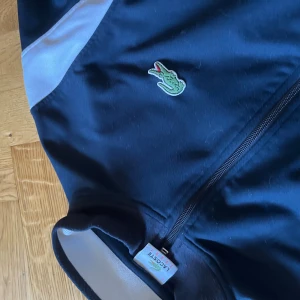 Blå tröja från Lacoste - Snygg svart tröja från Lacoste med dragkedja framtill, broderad logga på bröstet och stor Lacoste-text på ryggen. Jackan har vita detaljer på axlarna och är tillverkad i polyester. Perfekt för dig som gillar sportig och clean stil. Säljs i rent och snyggt skick, vid frågor är det bara att höra av sig!