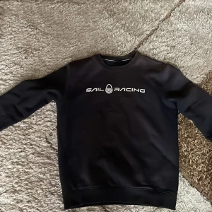 Svart Sail Racing sweatshirt M - Svart sweatshirt från Sail Racing i storlek M. Tröjan har en stilren design med Sail Racing-logga tryckt på bröstet och en mindre logga i nacken. Rund hals och långa ärmar, tillverkad i mjuk bomullsmix som är skön mot huden.