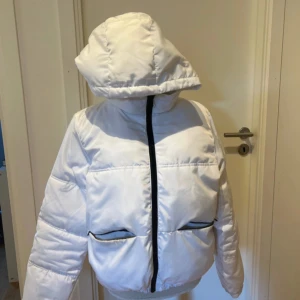 Vit pufferjacka med huva och svarta detaljer - Snygg vit pufferjacka med stor huva och svarta dragkedjor. Jackan har två stora fickor framtill och är perfekt för kalla dagar. Den är vadderad och har en modern, lite oversized passform som ger en cool look.lite fläck med fickan