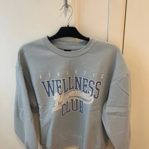 Ljusblå sweatshirt Wellness Movement Club - Ljusblå sweatshirt med trycket 'Wellness Movement Club' i vitt och blått framtill. Tröjan har rund halsringning, ribbade muddar och en avslappnad passform. Perfekt för en chill och sportig look.