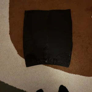 Svart jeanskjol från Vero Moda - Svart jeanskjol från Vero Moda i klassisk femficksmodell. Kjolen är rak och slutar ovanför knäna, med knapp och dragkedja framtill samt bakfickor. Perfekt för dig som gillar en enkel och stilren look.