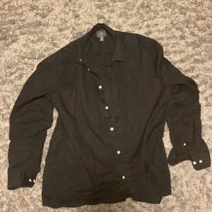 Svart skjorta från H&M med knappar - Svart skjorta från H&M i storlek L med klassisk krage och vita knappar framtill. Skjortan har lång ärm med knapp vid manschetten och är tillverkad i mjuk bomull. Perfekt för dig som gillar stilrena och enkla plagg.