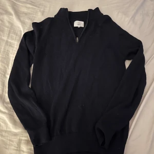 Mörkblå stickad tröja från Bläck - Snygg mörkblå stickad tröja från Bläck i storlek S. Tröjan har en half zip-dragkedja vid halsen och är långärmad med ribbade muddar. Perfekt för dig som gillar enkel och stilren design med lite sportig touch.