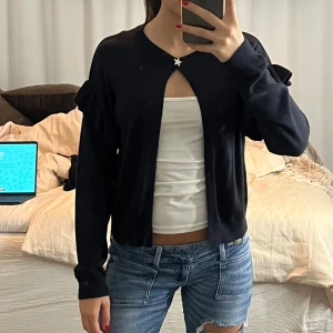 Svart kofta med volangdetaljer - Svart kofta med lång ärm och volangdetaljer på axlarna. Koftan har en söt stjärnformad knapp upptill och är öppen framtill. Mjuk och tunn, perfekt att slänga över ett linne eller t-shirt. Enkel och stilren design som funkar året runt.