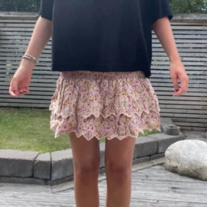 Blommig volangkjol med spetskant - Supersöt kort kjol med blommigt mönster i ljusa toner och volanger. Kjolen har en scallopad spetskant längst ner som ger en extra gullig touch. Perfekt för dig som gillar romantisk och feminin stil.