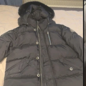 Svart dunjacka Moose Knuckles - Säljer en svart dunjacka från Moose Knuckles med fet pälskant på huvan och ikonisk logga på ärmen. Jackan har dragkedja, knappar och flera fickor. Broderad hockeypatch på insidan ger extra attityd. Perfekt för kalla dagar.