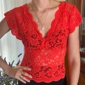 Röd spetsblus med v-ringning - Säljer en röd blus i spets med v-ringad hals och korta ärmar. Blusen har ett blommigt spetsmönster och vågiga kanter både vid halsringning och nederkant. Perfekt för dig som vill sticka ut med en färgstark och detaljerad topp.