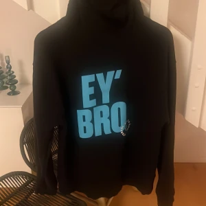 Ey Bro Hoodie  - Svart hoodie från EY'BRO med stor blå text 'EY' BRO' på ryggen. Huvan har coolt mönstrat innerfoder med vita motiv och snörning framtill. Tröjan är långärmad och har en klassisk passform. Skick-8,5/10.                                                          