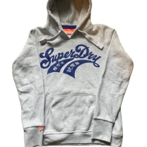 Grå Superdry hoodie med blå logga - Snygg grå hoodie från Superdry med stor blå logga och japanska tecken på bröstet. Tröjan har huva med snörning och en stor magficka. Perfekt för dig som gillar streetwear och vill ha en bekväm och stilren look.