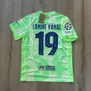 Barcelona Lamine Yamal fotbollströja - Säljer en officiell FC Barcelona fotbollströja från Nike med Lamine Yamal och nummer 19 på ryggen. Tröjan är ljusgrön med mönstrade detaljer, korta ärmar och tryckta loggor från Spotify, UNHCR och UEFA Foundation. Materialet är lätt och andas, perfekt för match eller träning.