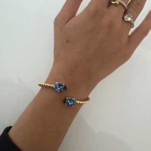 Guldfärgat armband med blå stenar - Säljer den för Halva priset!!                               Snyggt guldfärgat armband med tvinnad design och två stora droppformade blå stenar i varje ände. Armbandet är öppet och ger en lyxig känsla med sina glittrande detaljer. Perfekt accessoar för att lyfta din outfit.