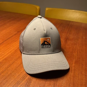 Grå Tierra keps med mesh och broderi - Snygg grå keps från Tierra med böjd skärm och mesh på baksidan för extra ventilation. Framtill finns ett broderat Tierra-märke med bergmotiv i svart och orange. Justerbar snapback för perfekt passform. Perfekt för dig som gillar outdoor och streetstyle. Aldrig använd så det är grymt skick 