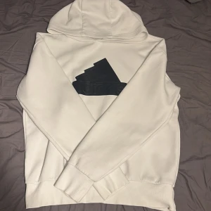 Vit hoodie från Adidas med svart logga - Vit hoodie från Adidas med stor svart 3D-logga på bröstet. Klassisk modell med luva och. Perfekt för dig som gillar sportig och clean stil. Tillverkad i mjuk bomullsblandning för extra komfort.