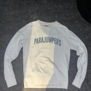 Vit Parajumpers tröja - Vit långärmad fleece-tröja från Parajumpers med tryckt logga i grått framtill och orange detalj på insidan av nederkanten. Tröjan har ribbade muddar och en patch på ena ärmen. Perfekt för dig som gillar clean och sportig stil.