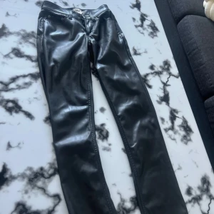 Svarta byxor i skinnimitation - Säljer ett par svarta byxor i skinnimitation med klassisk femficksdesign och raka ben. Byxorna har normal passform och midja, samt dragkedja och knapp framtill. Perfekta för dig som vill ha en edgy look.