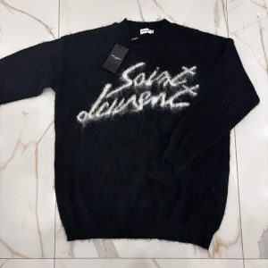 Saint Laurent stickad tröja - Svart stickad tröja från Saint Laurent med vit, fluffig logotyp framtill. Tröjan har rund halsringning och långa ärmar. Den är gjord i ett mjukt, lurvigt material som ger en cool och lyxig känsla. Perfekt för dig som vill sticka ut från mängden. Passformen på den är s/m.