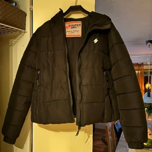 Svart pufferjacka från Superdry - Svart pufferjacka från Superdry Sports Puffer Series. Jackan har huva, dragkedja framtill och två sidofickor med dragkedja. Klassisk quiltad look och sportig vibe, perfekt för kalla dagar. Snygg och enkel att matcha med det mesta.