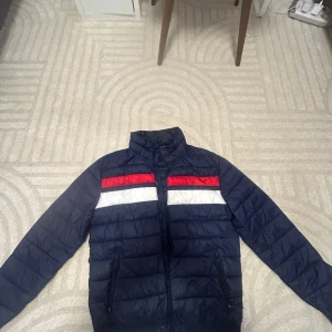 Mörkblå pufferjacka med röd och vit detalj - Säljer en mörkblå pufferjacka med horisontella röda och vita ränder över bröstet och ryggen. Jackan har hög krage, dragkedja framtill och två fickor med dragkedja. Perfekt för kyliga dagar och har en sportig vibe.